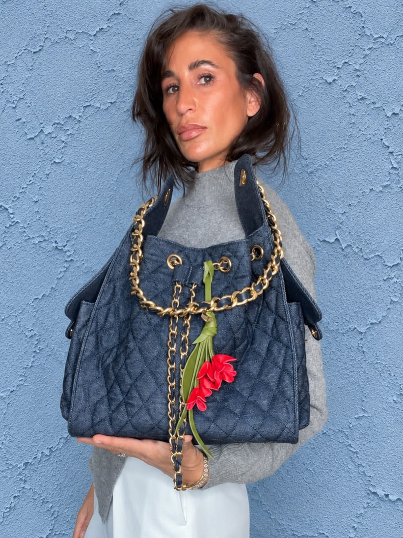 Renee, founder and designer of BEACH HAUS, holding a quilted denim bucket bag with a red flower charm against a blue wall